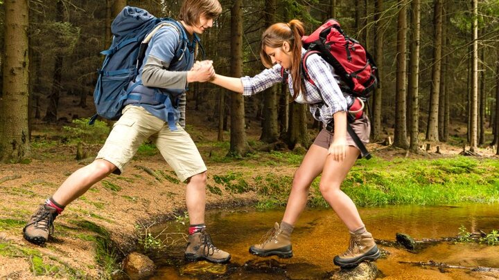 Stock photo of a boy helping a girl cross a stream while hiking.