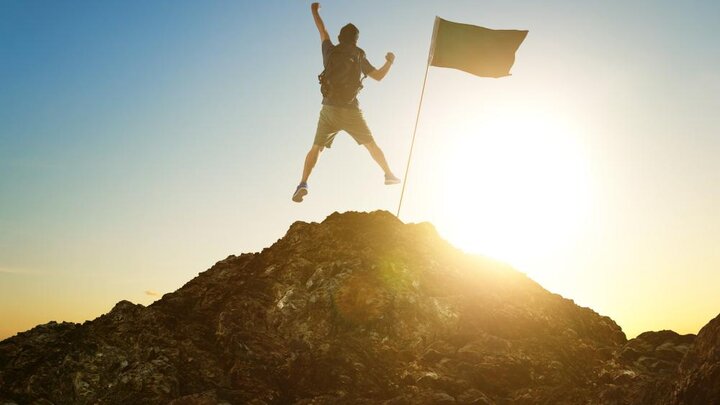 Success, leadership, achievement and people concept - stock photo of silhouette of young guy with flag on mountain top over sky and sun light background.