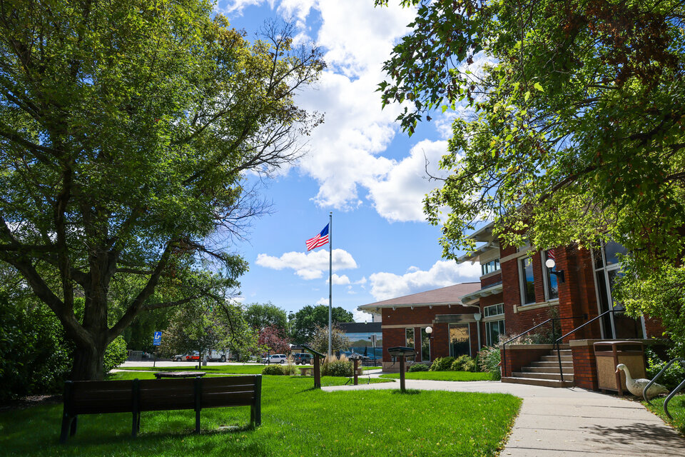 Picture of a walking trail leading past the Tekamah community library, a red brick building, on the right, with a park bench on the left, and an American flag on a pole in the background middle. Green trees border the picture on either saide, and the blue sky has clouds in it.