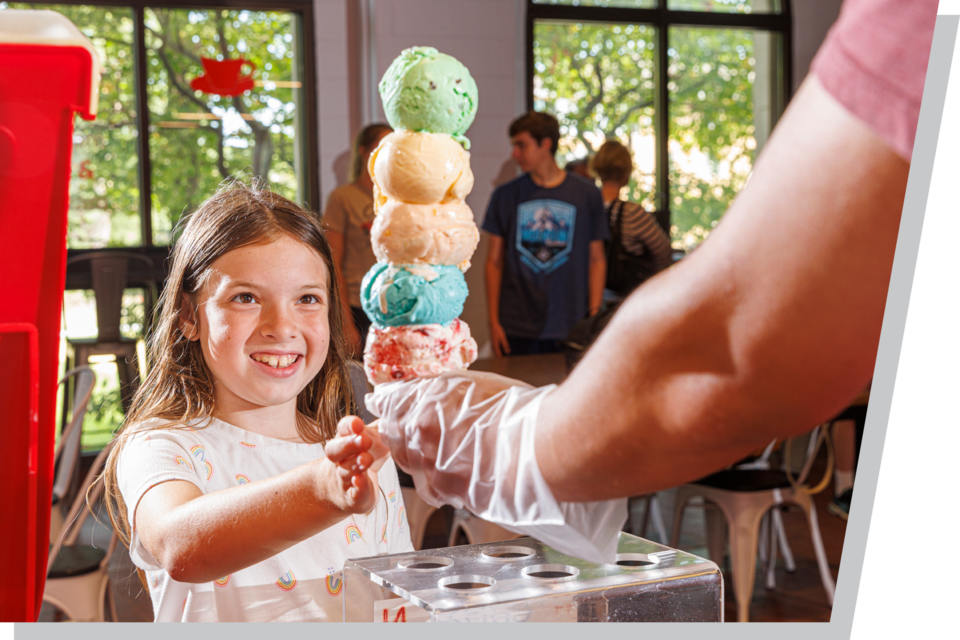 Child receiving a stacked ice cream cone from a vendor in a bright indoor setting.