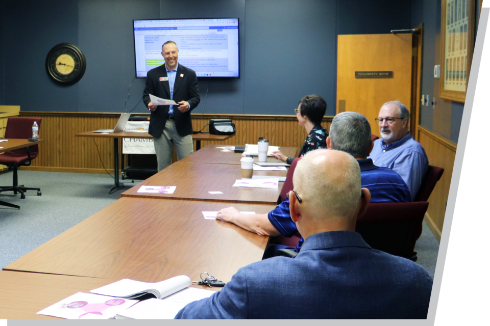 A man stands at the front of a board room, in front of a screen, presenting to a room full of business professionals.