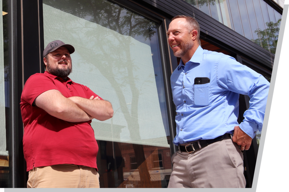 Two men stand in front of a store front in a rural downtown area.