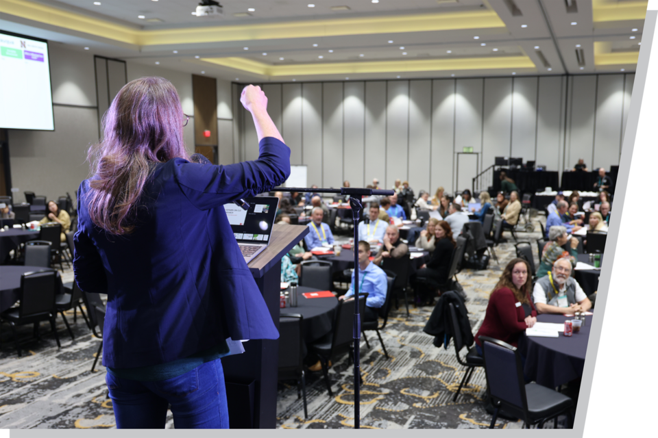 A woman stands at a podium, her back to the camera, presenting to a conference room full of people.