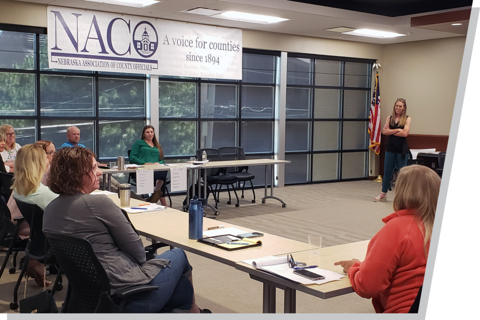 A woman stands behind a podium, presenting to a classroom of adult leaners.