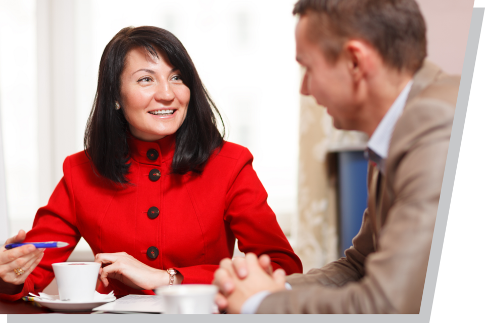 Business meeting with two people seated, one in a red jacket holding a pen, and coffee cups on the table.