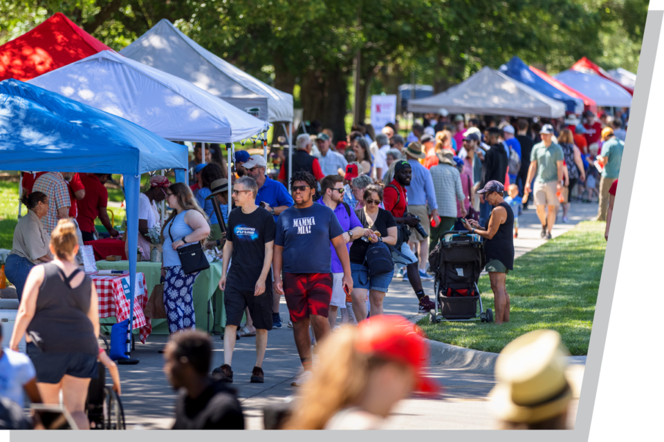 Crowd walking through outdoor market with colorful tents and green trees.