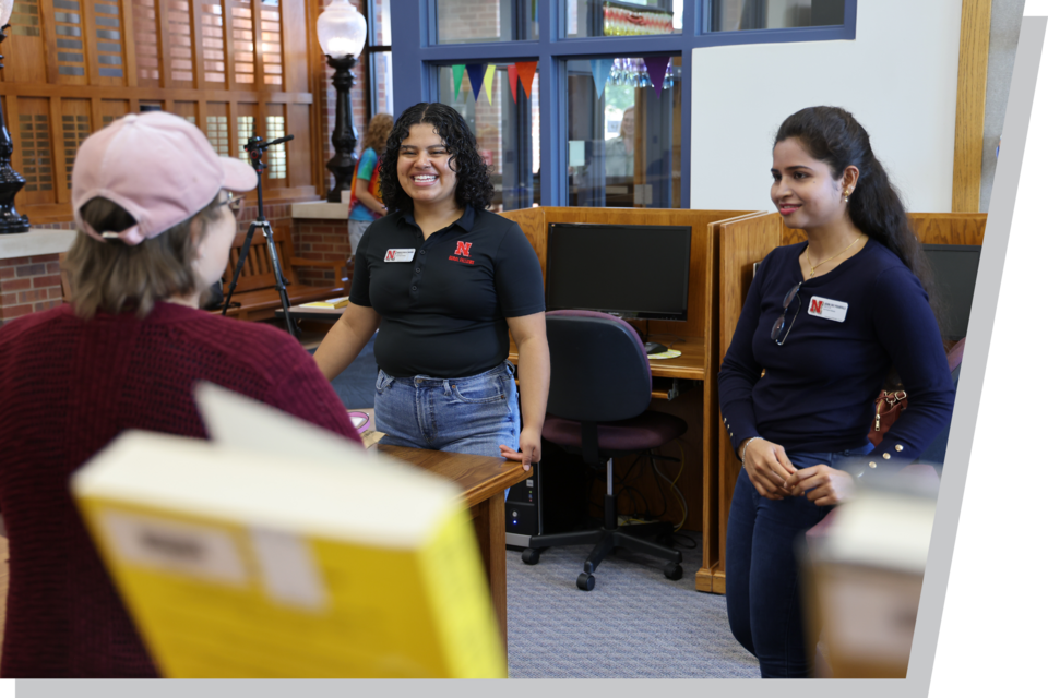 Two people with name tags converse with another person in a library setting near desks and computers.
