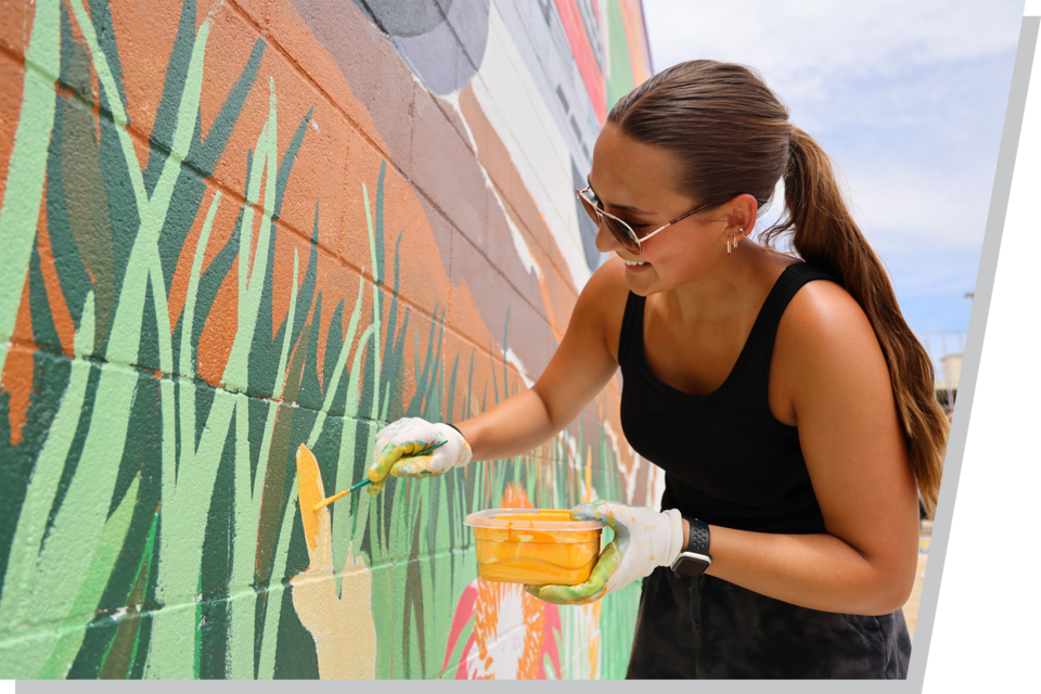 A woman holding a yellow paint can and painting a flower on a mural.