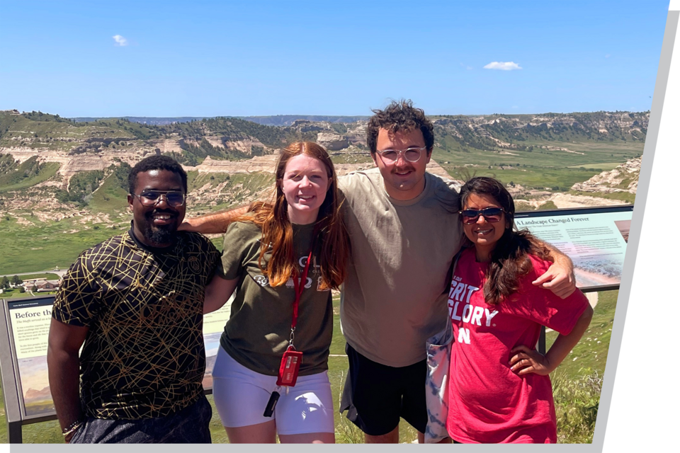 Four college-age young adults, two men and two men, stand arm in arm in front of the Scottsbluff momument in Nebraska.