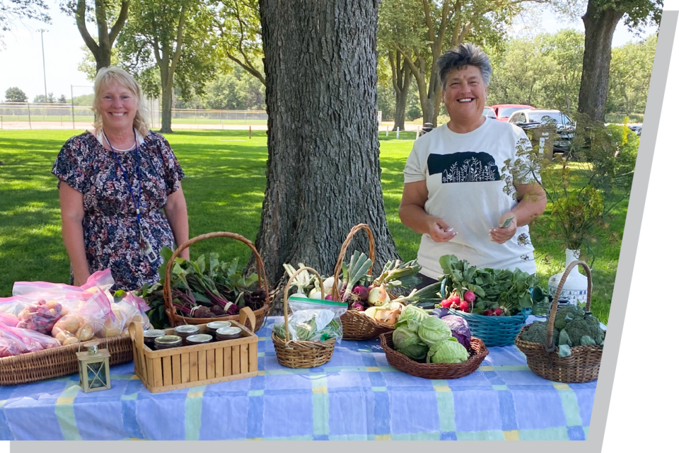 Farmers market stand with vegetables and goods displayed on a table under trees.