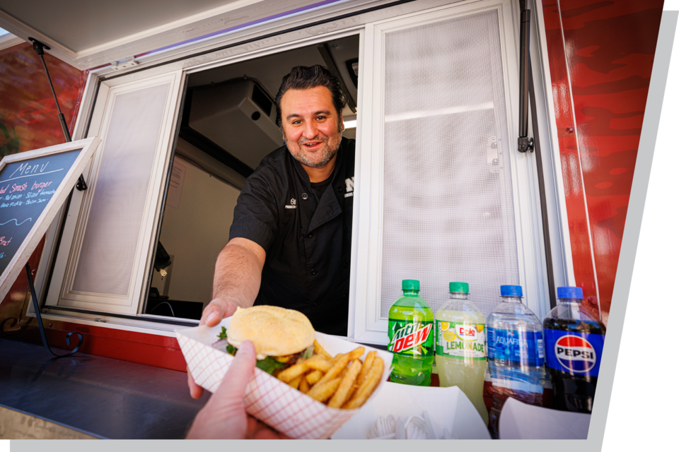 Food truck worker handing out a burger and fries tray; assorted drinks displayed beside the window.