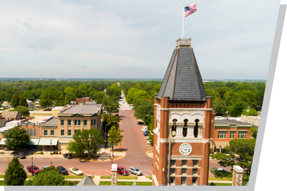 Brick clock tower with an American flag surrounded by a small-town landscape and greenery.